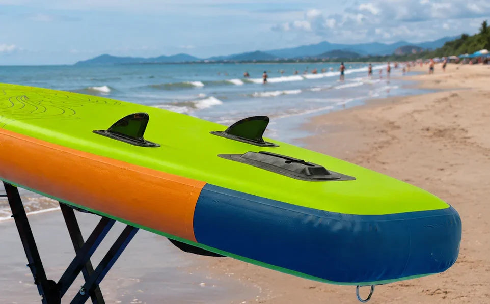 Bright green, orange, and blue inflatable SUP paddle board with fins on beach near ocean waves